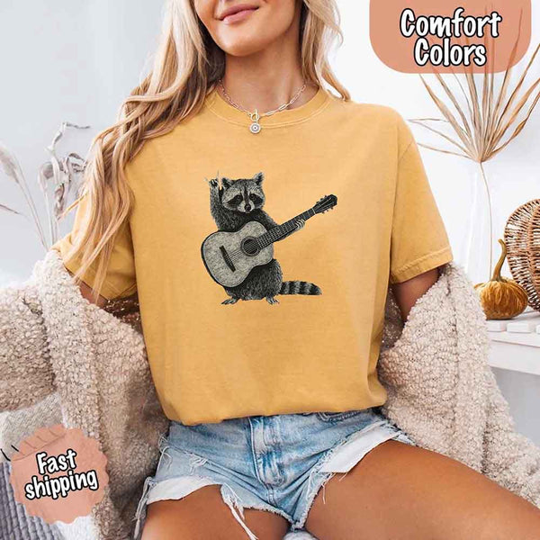 May include a woman wearing a mustard Comfort Colors T-shirt featuring a graphic of a raccoon playing a guitar, styled for a quirky and fun animal lover look.