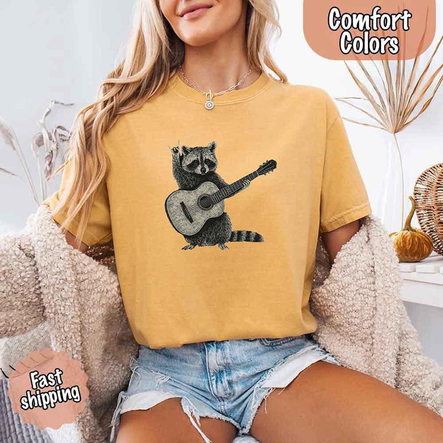 May include a woman wearing a mustard Comfort Colors T-shirt featuring a graphic of a raccoon playing a guitar, styled for a quirky and fun animal lover look.