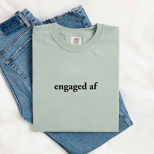 May include a folded bay Comfort Colors T-shirt with “engaged AF” printed in black, laid over denim jeans for a playful and celebratory look.