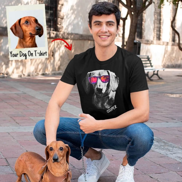 Man wearing a black t-shirt with a dog graphic and a dachshund on a leash outdoors.