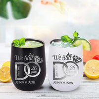 Two tumblers with 'We Still Do' engraving on a wooden surface with fruit and plant in the background.
