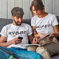 Man and woman sitting on a couch wearing 'HUBBY' and 'WIFEY' shirts with establishment years.