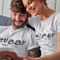 Couple wearing matching t-shirts with 'Hubby' and 'Wifey' text, smiling and looking at a device.