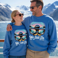 Smiling couple in matching custom blue sweatshirts reading 'We Still Do,' scenic mountains background