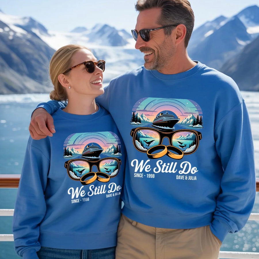 Smiling couple in matching custom blue sweatshirts reading 'We Still Do,' scenic mountains background