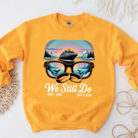 Yellow sweatshirt with cruise ship, mountains, sunglasses, wedding rings, and 'We Still Do' text