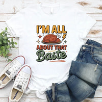 White t-shirt with 'I'm All About That Baste' graphic, paired with jeans and sneakers on a wooden surface.