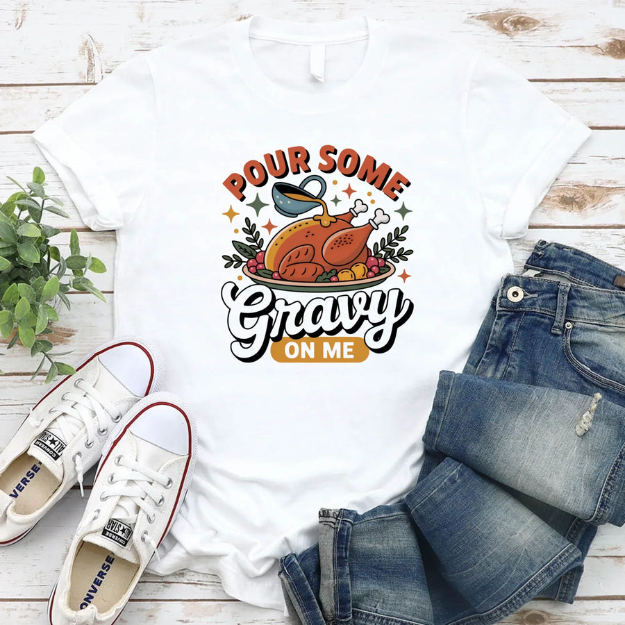 White t-shirt with 'Pour Some Gravy On Me' graphic, paired with jeans and sneakers on a wooden surface.
