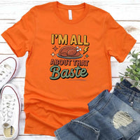 Orange t-shirt with 'I'm all about that baste' text and graphic, placed on a wooden surface with jeans and sneakers.