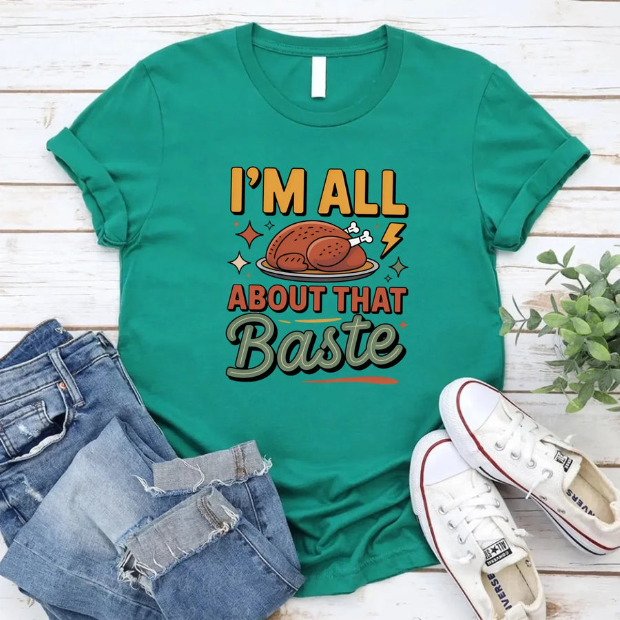 Green t-shirt with 'I'm All About That Baste' text and graphic, paired with jeans and sneakers on a wooden surface.