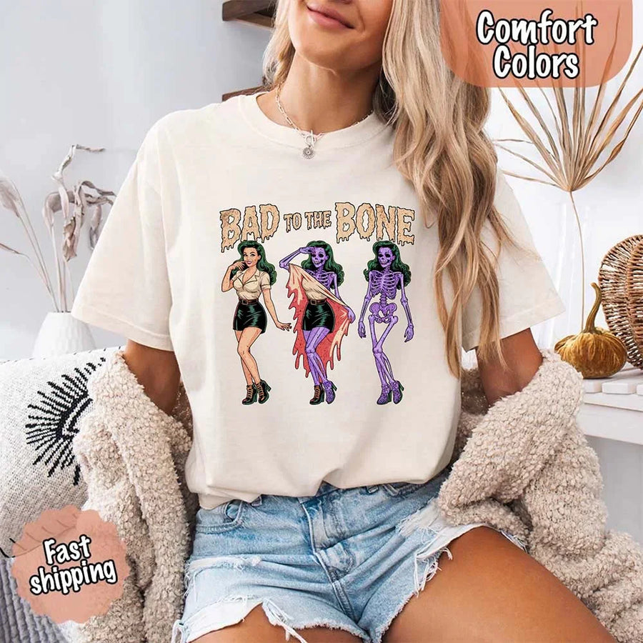 May include Bad to the Bone graphic with retro pin-up girl transforming into purple skeleton artwork – vintage Halloween shirt.