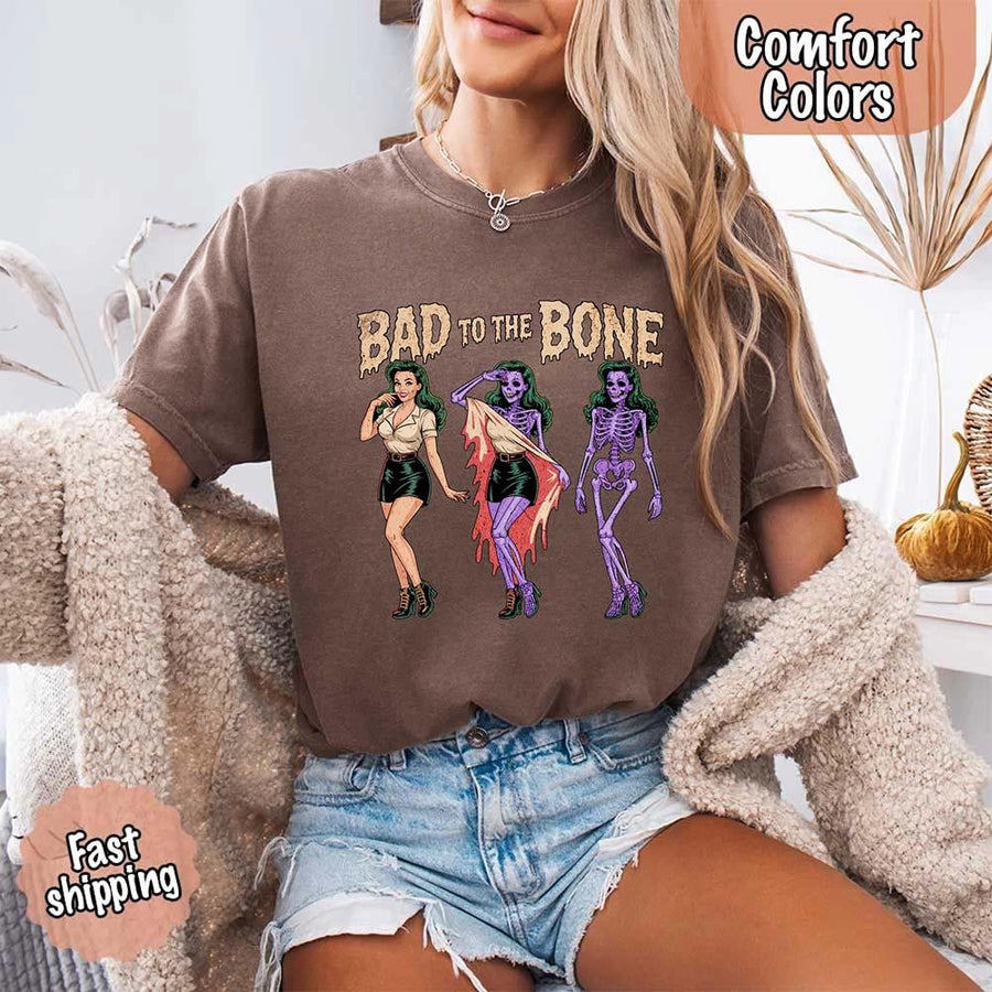 May include Bad to the Bone graphic with retro pin-up girl transforming into purple skeleton artwork – vintage Halloween shirt.