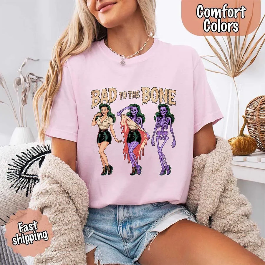 May include Bad to the Bone graphic with retro pin-up girl transforming into purple skeleton artwork – vintage Halloween shirt.