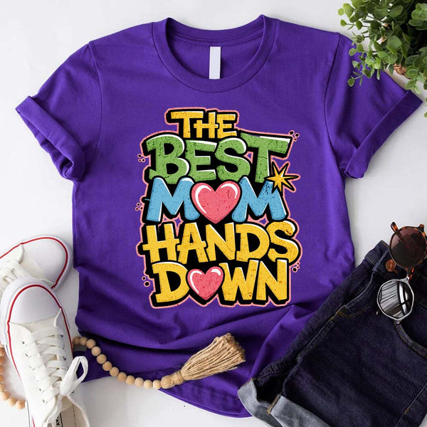 A woman sitting on a couch wearing a white t-shirt with the text 'THE BEST MOM HANDS DOWN' printed on it.