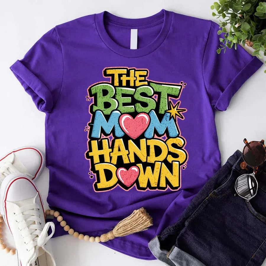 A woman sitting on a couch wearing a white t-shirt with the text 'THE BEST MOM HANDS DOWN' printed on it.