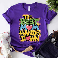 A woman sitting on a couch wearing a white t-shirt with the text 'THE BEST MOM HANDS DOWN' printed on it.