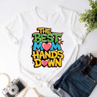 A woman sitting on a couch wearing a white t-shirt with the text 'THE BEST MOM HANDS DOWN' printed on it.