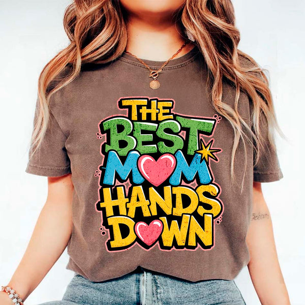A woman sitting on a couch wearing a white t-shirt with the text 'THE BEST MOM HANDS DOWN' printed on it.