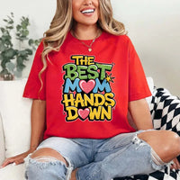A woman sitting on a couch wearing a white t-shirt with the text 'THE BEST MOM HANDS DOWN' printed on it.
