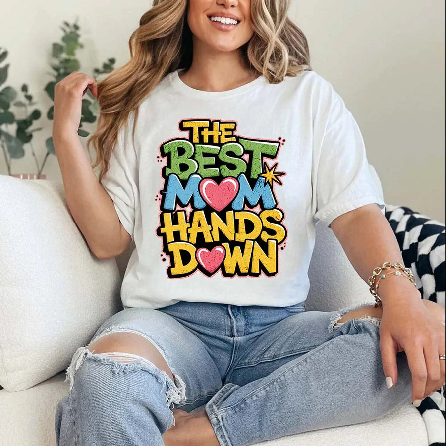 A woman sitting on a couch wearing a white t-shirt with the text 'THE BEST MOM HANDS DOWN' printed on it.