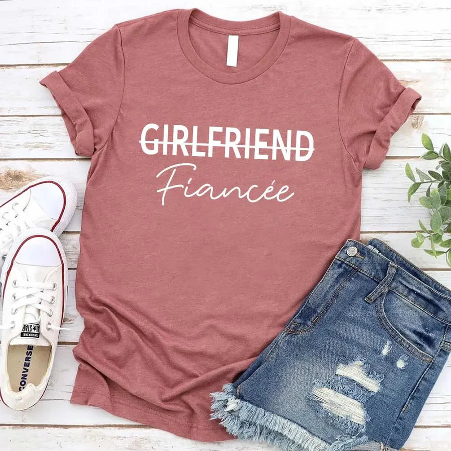 Black t-shirt with 'GIRLFRIEND Fiançée' text, paired with jeans and sneakers on a wooden surface.