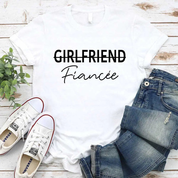 Black t-shirt with 'GIRLFRIEND Fiançée' text, paired with jeans and sneakers on a wooden surface.