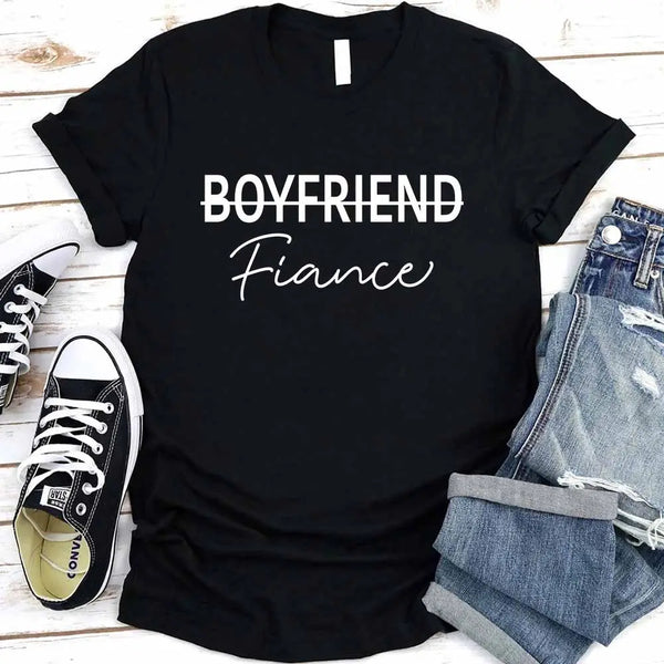 Black t-shirt with 'BOYFRIEND Fiançée' text, paired with jeans and sneakers on a wooden surface.