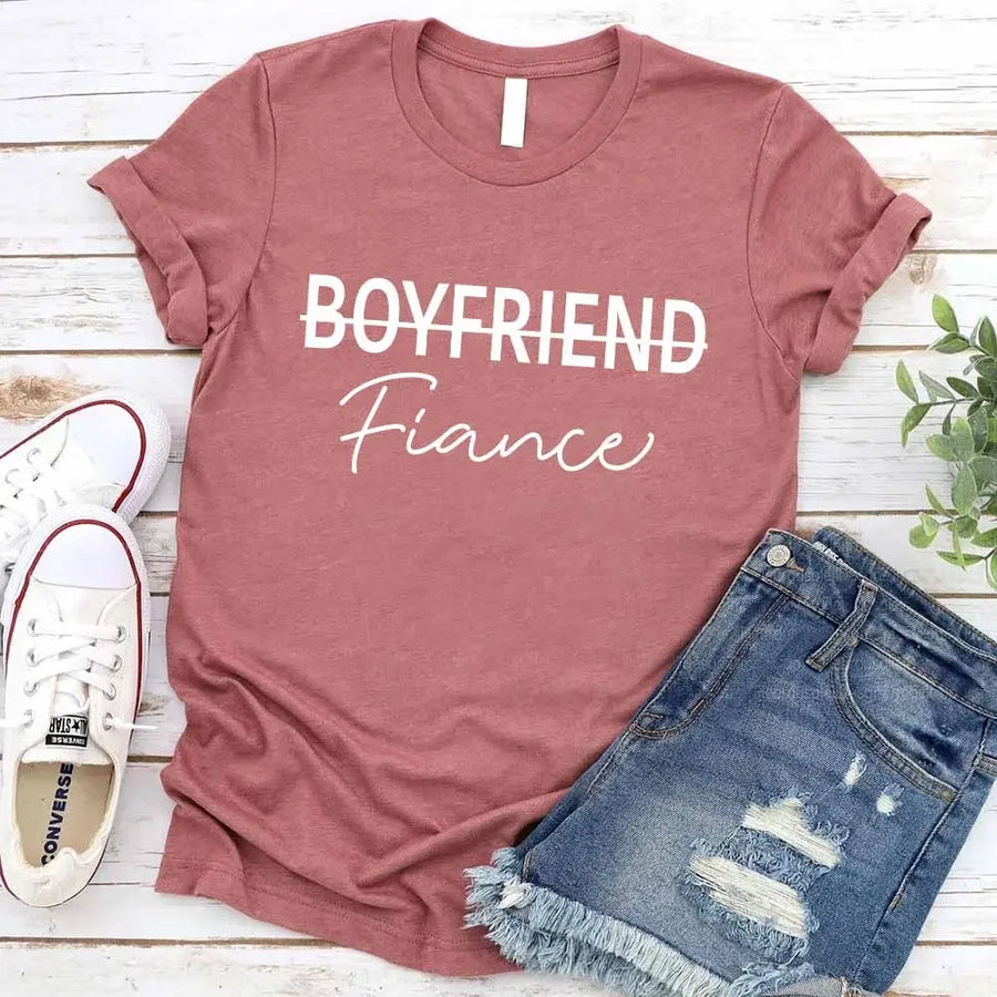 Black t-shirt with 'BOYFRIEND Fiançée' text, paired with jeans and sneakers on a wooden surface.