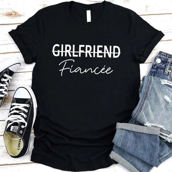 Black t-shirt with 'GIRLFRIEND Fiançée' text, paired with jeans and sneakers on a wooden surface.