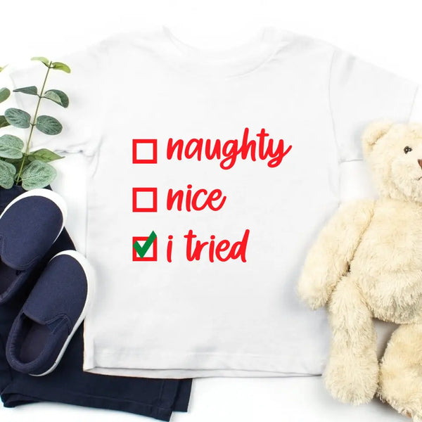 May include a white Christmas t-shirt displayed flat with festive decorations and gift boxes around it. The shirt features white text that reads “naughty,” “nice,” and “i tried,” with checkboxes next to each option. The “i tried” box is marked with a green check, creating a playful and humorous holiday design.