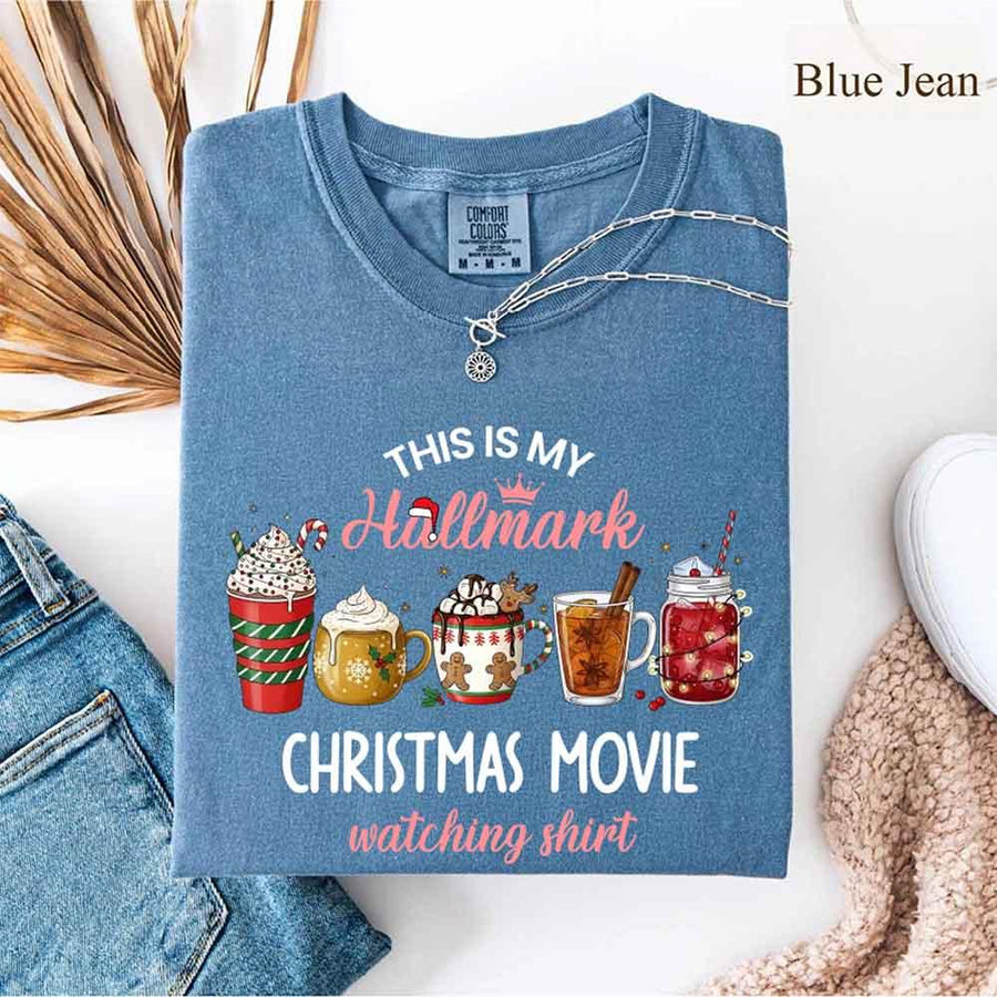 blue jean Comfort Colors t-shirt with a festive Christmas design. The shirt features the phrase “This Is My Hallmark Christmas Movie Watching Shirt” in playful fonts, with “Hallmark” written in pink cursive topped with a small crown and Santa hat. Below the text are five illustrated holiday beverages: a red and green striped cup with whipped cream and candy cane, a golden mug with whipped cream, a white mug with gingerbread men and holly, a tall glass of tea with cinnamon stick and star anise, and a maso
