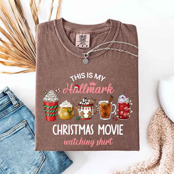 Espresso Comfort Colors t-shirt with a festive Christmas design. The shirt features the phrase “This Is My Hallmark Christmas Movie Watching Shirt” in playful fonts, with “Hallmark” written in pink cursive topped with a small crown and Santa hat. Below the text are five illustrated holiday beverages: a red and green striped cup with whipped cream and candy cane, a golden mug with whipped cream, a white mug with gingerbread men and holly, a tall glass of tea with cinnamon stick and star anise, and a maso
