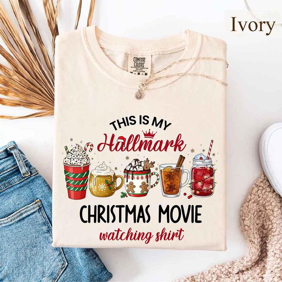 Ivory Comfort Colors t-shirt with a festive Christmas design. The shirt features the phrase “This Is My Hallmark Christmas Movie Watching Shirt” in playful fonts, with “Hallmark” written in pink cursive topped with a small crown and Santa hat. Below the text are five illustrated holiday beverages: a red and green striped cup with whipped cream and candy cane, a golden mug with whipped cream, a white mug with gingerbread men and holly, a tall glass of tea with cinnamon stick and star anise, and a maso