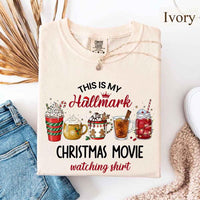 Ivory Comfort Colors t-shirt with a festive Christmas design. The shirt features the phrase “This Is My Hallmark Christmas Movie Watching Shirt” in playful fonts, with “Hallmark” written in pink cursive topped with a small crown and Santa hat. Below the text are five illustrated holiday beverages: a red and green striped cup with whipped cream and candy cane, a golden mug with whipped cream, a white mug with gingerbread men and holly, a tall glass of tea with cinnamon stick and star anise, and a maso