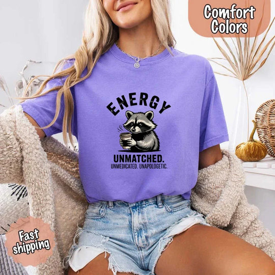 May include a black and white raccoon illustration with a tired, unbothered expression, holding a coffee cup, and bold text that reads “ENERGY UNMATCHED. UNMEDICATED. UNAPOLOGETIC.” conveying raw, unapologetic humor and mood.