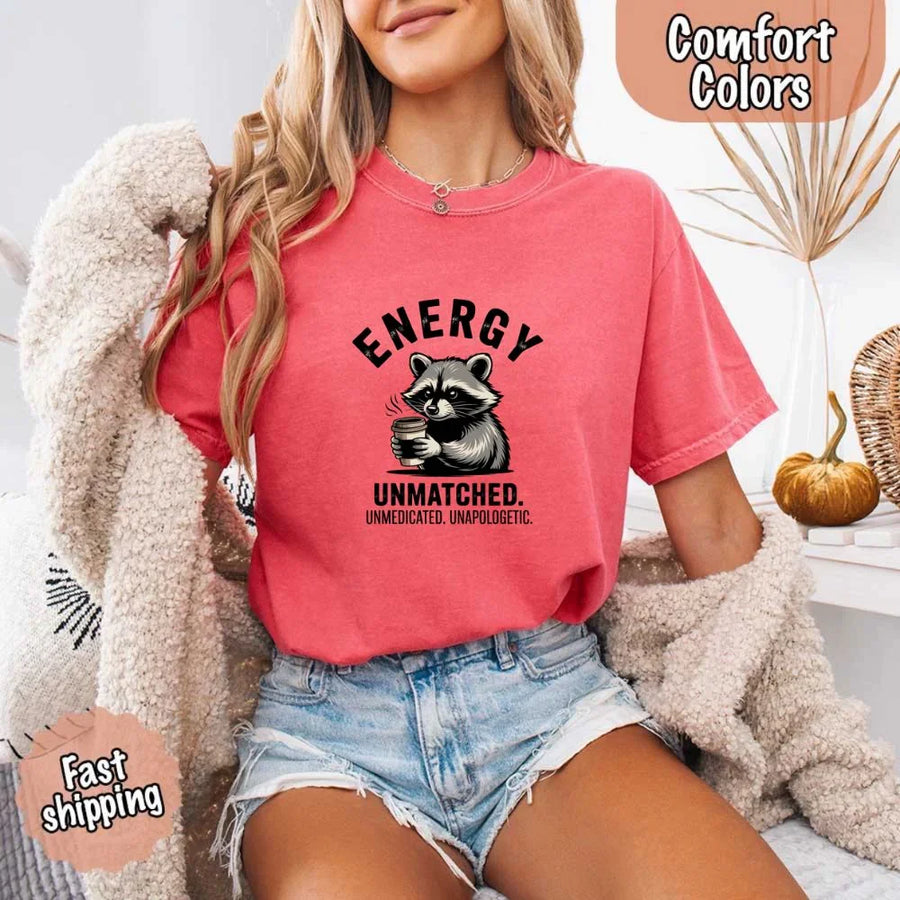 May include a black and white raccoon illustration with a tired, unbothered expression, holding a coffee cup, and bold text that reads “ENERGY UNMATCHED. UNMEDICATED. UNAPOLOGETIC.” conveying raw, unapologetic humor and mood.