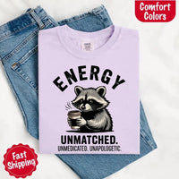 May include a black and white raccoon illustration with a tired, unbothered expression, holding a coffee cup, and bold text that reads “ENERGY UNMATCHED. UNMEDICATED. UNAPOLOGETIC.” conveying raw, unapologetic humor and mood.