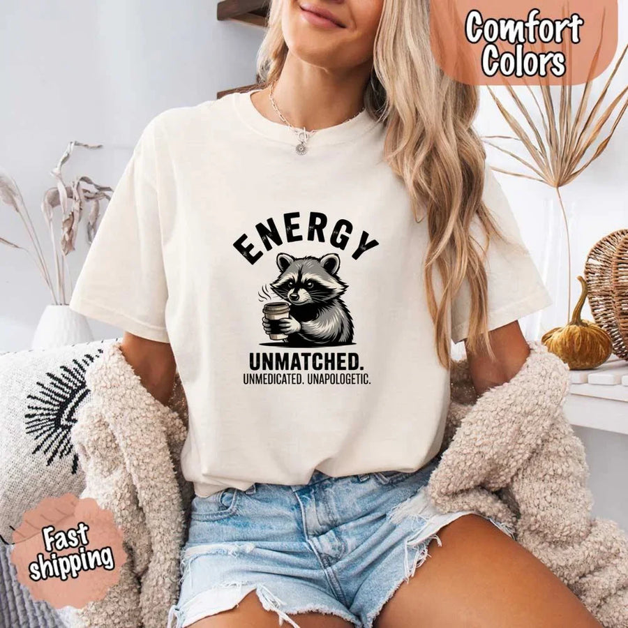 May include a black and white raccoon illustration with a tired, unbothered expression, holding a coffee cup, and bold text that reads “ENERGY UNMATCHED. UNMEDICATED. UNAPOLOGETIC.” conveying raw, unapologetic humor and mood.