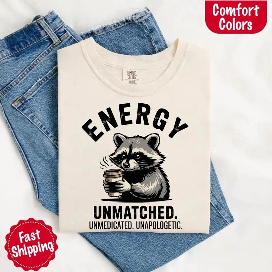 May include a black and white raccoon illustration with a tired, unbothered expression, holding a coffee cup, and bold text that reads “ENERGY UNMATCHED. UNMEDICATED. UNAPOLOGETIC.” conveying raw, unapologetic humor and mood.