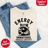 May include a black and white raccoon illustration with a tired, unbothered expression, holding a coffee cup, and bold text that reads “ENERGY UNMATCHED. UNMEDICATED. UNAPOLOGETIC.” conveying raw, unapologetic humor and mood.