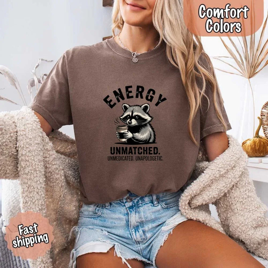 May include a black and white raccoon illustration with a tired, unbothered expression, holding a coffee cup, and bold text that reads “ENERGY UNMATCHED. UNMEDICATED. UNAPOLOGETIC.” conveying raw, unapologetic humor and mood.