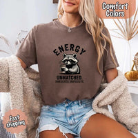 May include a black and white raccoon illustration with a tired, unbothered expression, holding a coffee cup, and bold text that reads “ENERGY UNMATCHED. UNMEDICATED. UNAPOLOGETIC.” conveying raw, unapologetic humor and mood.