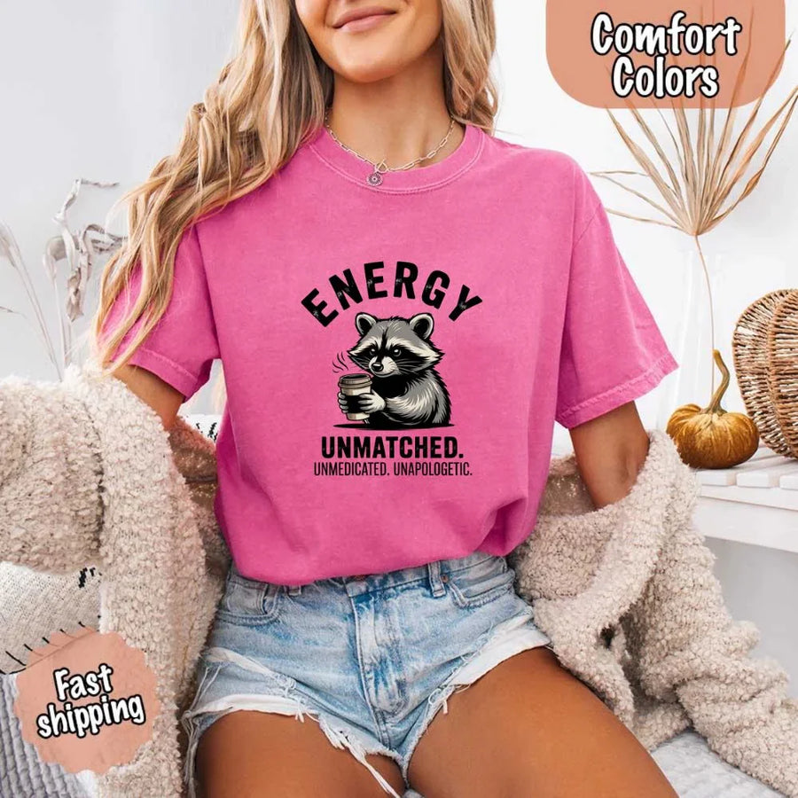May include a black and white raccoon illustration with a tired, unbothered expression, holding a coffee cup, and bold text that reads “ENERGY UNMATCHED. UNMEDICATED. UNAPOLOGETIC.” conveying raw, unapologetic humor and mood.