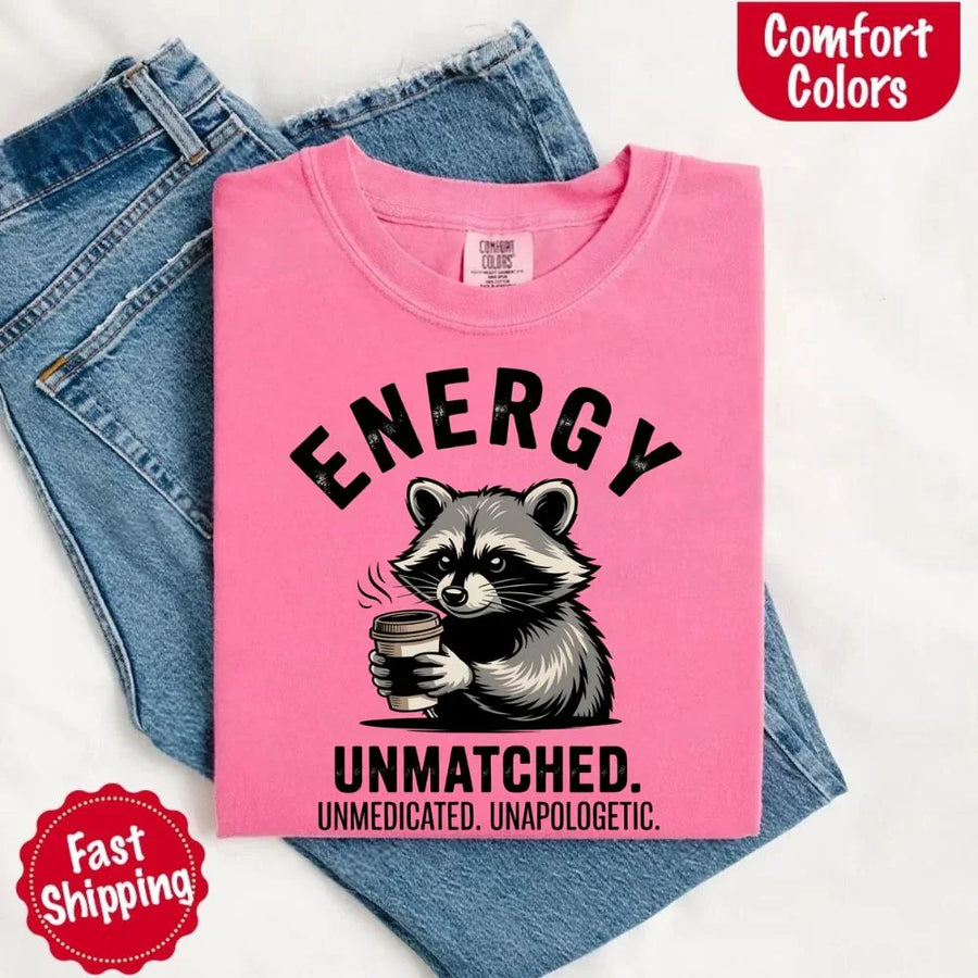 May include a black and white raccoon illustration with a tired, unbothered expression, holding a coffee cup, and bold text that reads “ENERGY UNMATCHED. UNMEDICATED. UNAPOLOGETIC.” conveying raw, unapologetic humor and mood.