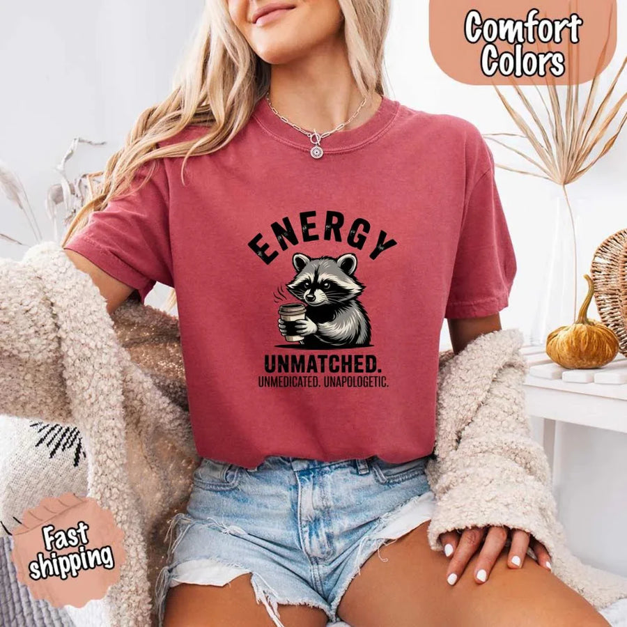 May include a black and white raccoon illustration with a tired, unbothered expression, holding a coffee cup, and bold text that reads “ENERGY UNMATCHED. UNMEDICATED. UNAPOLOGETIC.” conveying raw, unapologetic humor and mood.