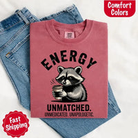 May include a black and white raccoon illustration with a tired, unbothered expression, holding a coffee cup, and bold text that reads “ENERGY UNMATCHED. UNMEDICATED. UNAPOLOGETIC.” conveying raw, unapologetic humor and mood.