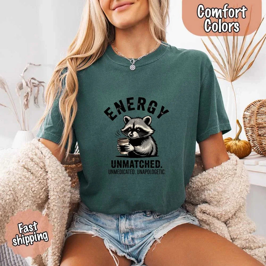 May include a black and white raccoon illustration with a tired, unbothered expression, holding a coffee cup, and bold text that reads “ENERGY UNMATCHED. UNMEDICATED. UNAPOLOGETIC.” conveying raw, unapologetic humor and mood.