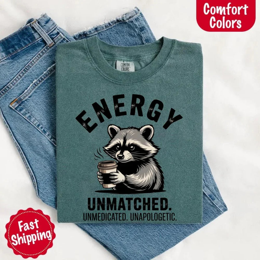 May include a black and white raccoon illustration with a tired, unbothered expression, holding a coffee cup, and bold text that reads “ENERGY UNMATCHED. UNMEDICATED. UNAPOLOGETIC.” conveying raw, unapologetic humor and mood.