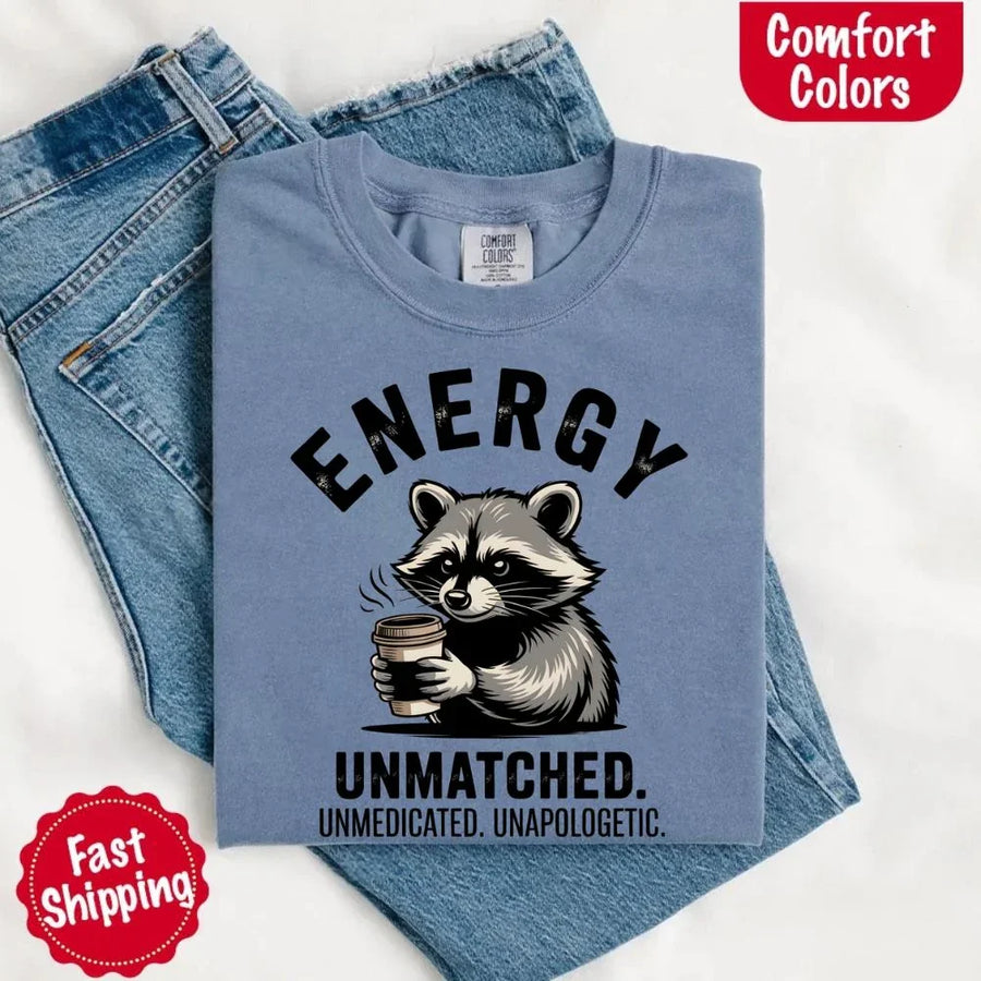 May include a black and white raccoon illustration with a tired, unbothered expression, holding a coffee cup, and bold text that reads “ENERGY UNMATCHED. UNMEDICATED. UNAPOLOGETIC.” conveying raw, unapologetic humor and mood.