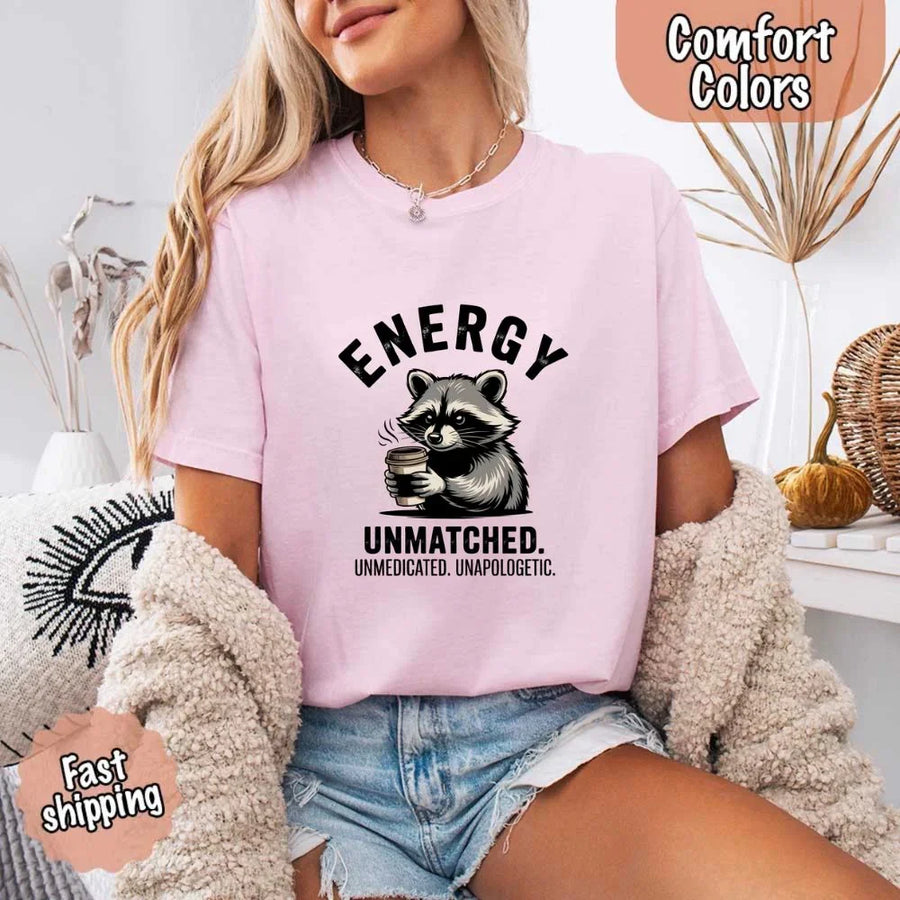 May include a black and white raccoon illustration with a tired, unbothered expression, holding a coffee cup, and bold text that reads “ENERGY UNMATCHED. UNMEDICATED. UNAPOLOGETIC.” conveying raw, unapologetic humor and mood.
